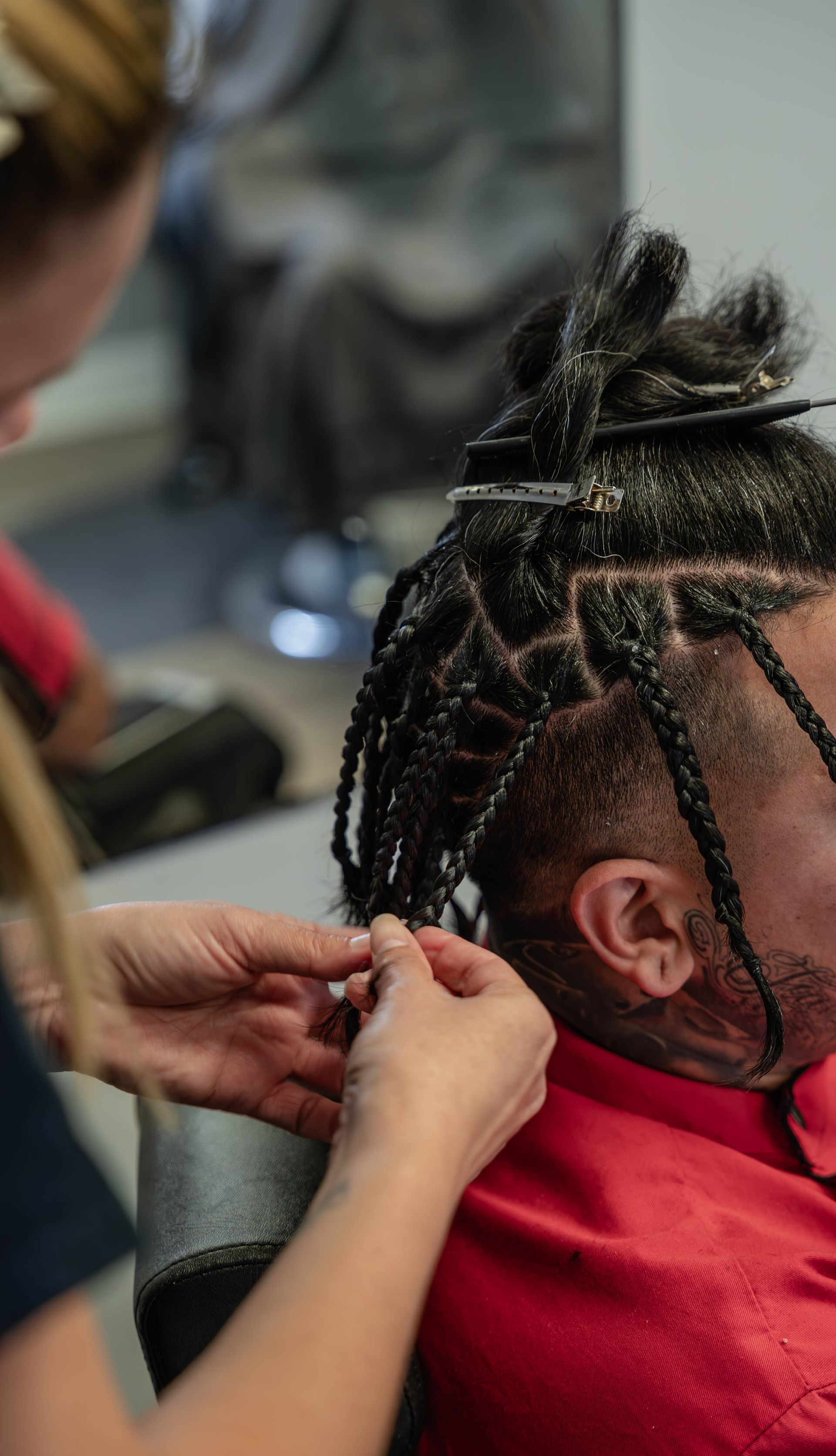 A stylist braids hair in small, box-section segments on a person wearing a red shirt.  The Studio Barbershop