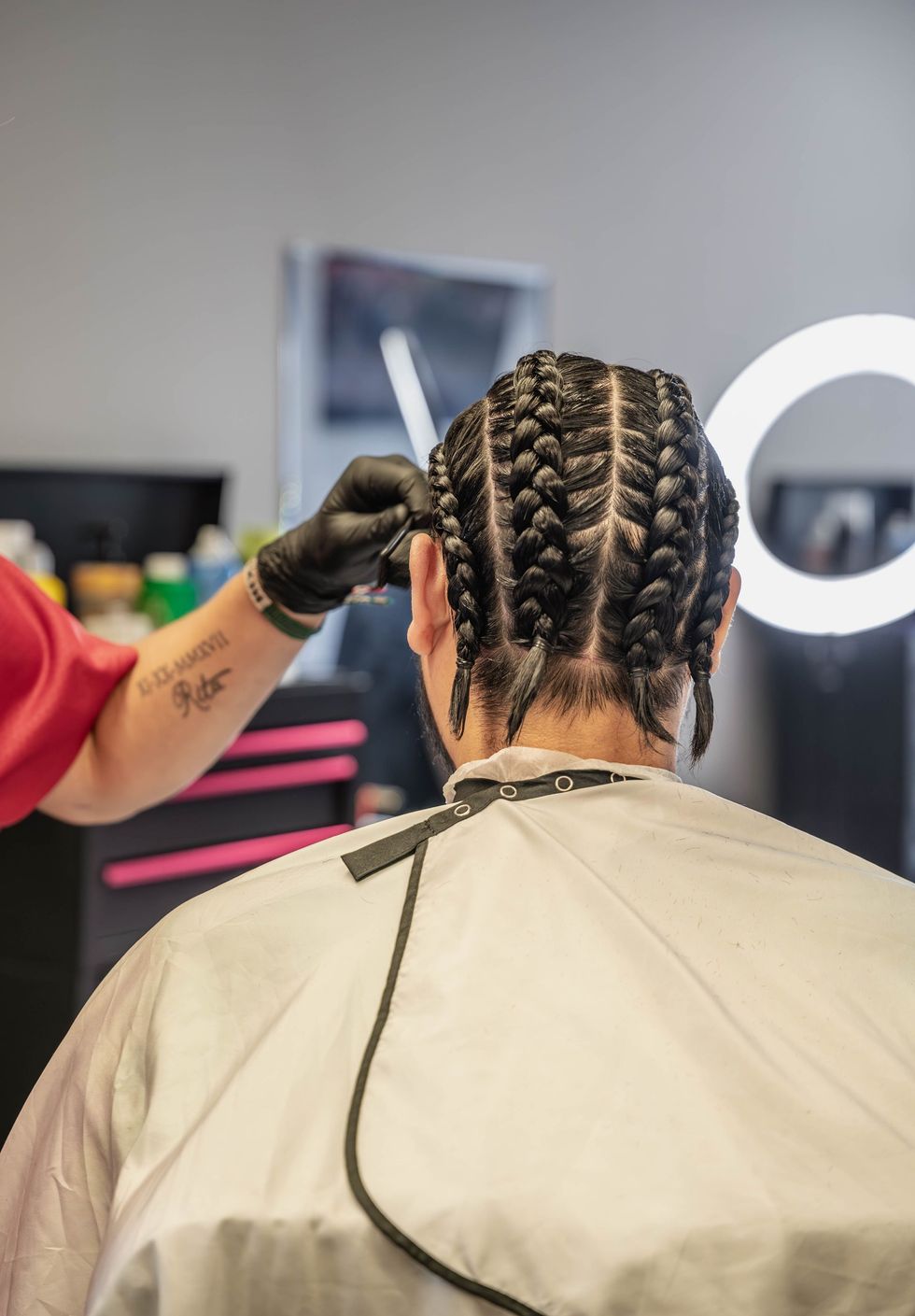 A barber wearing black gloves trims the back of a client's hair, which is styled in several neat cornrows.  The Studio Barbershop