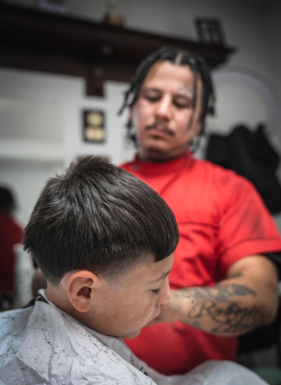 A barber in a red shirt cuts a person's hair in a salon, focusing closely on the client's fade.