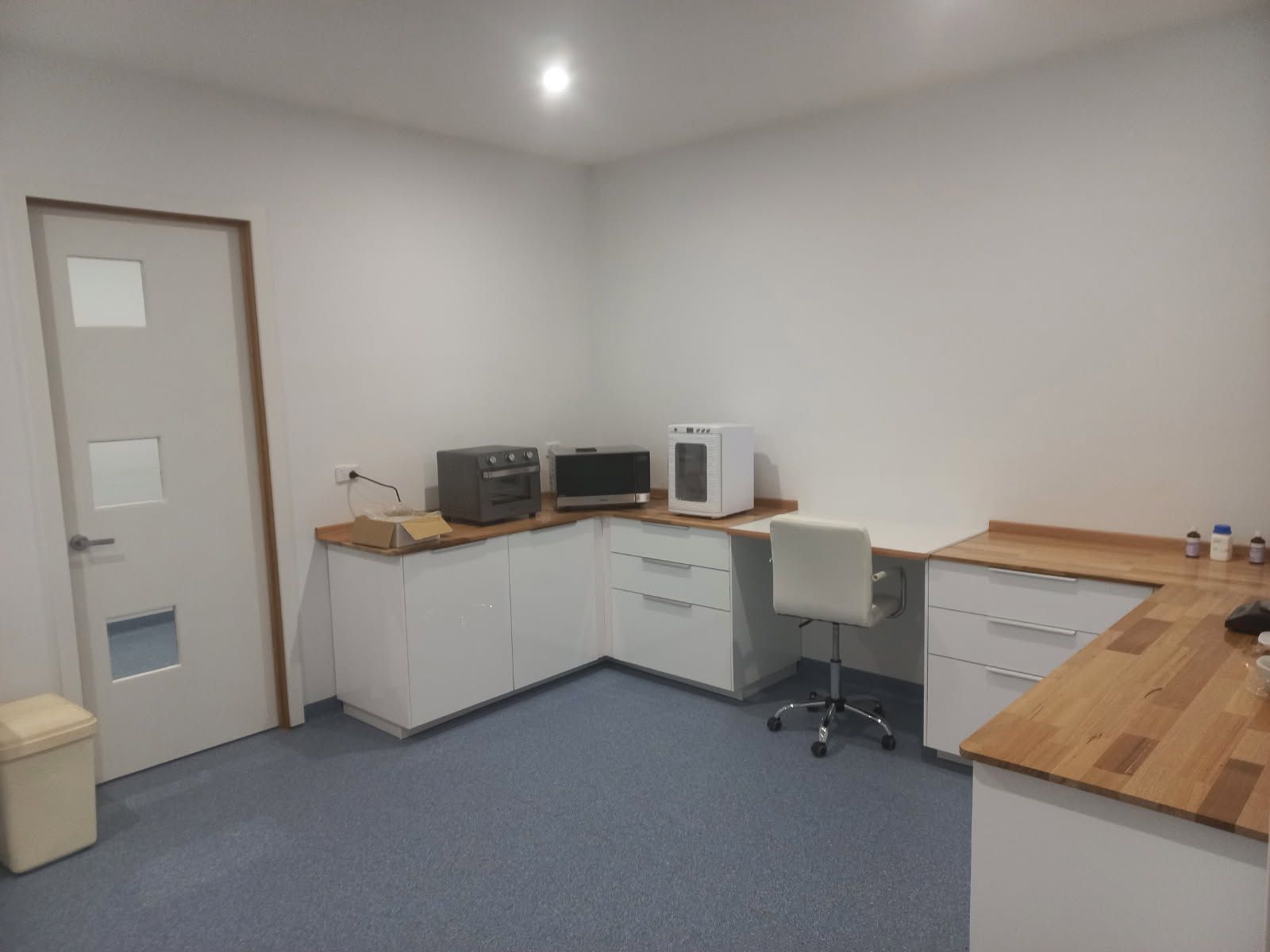 An empty, bright office kitchenette with white cabinets, wooden countertops, and a blue floor. 