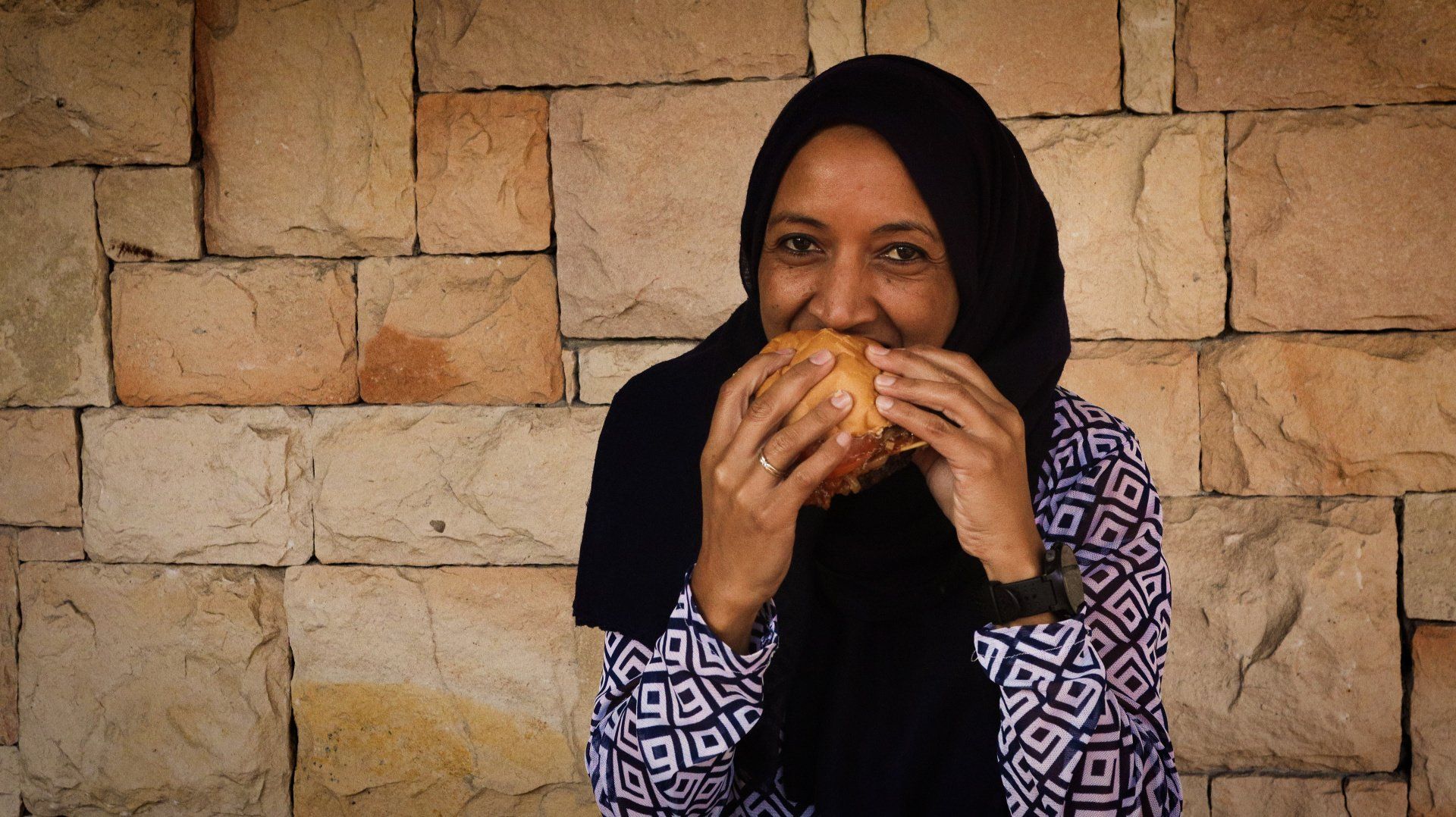 A woman in a hijab is eating a hamburger in front of a brick wall.