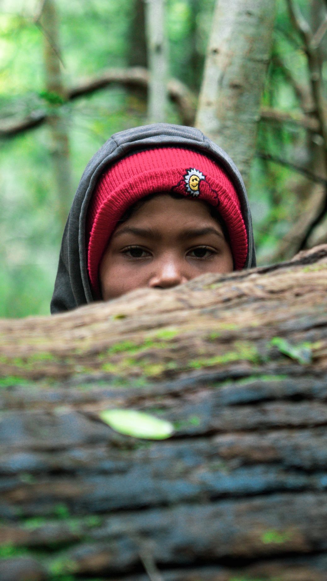 A young boy wearing a red beanie is peeking over a log in the woods.