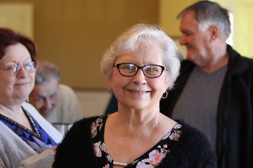 A woman wearing glasses is smiling in a room with other people.