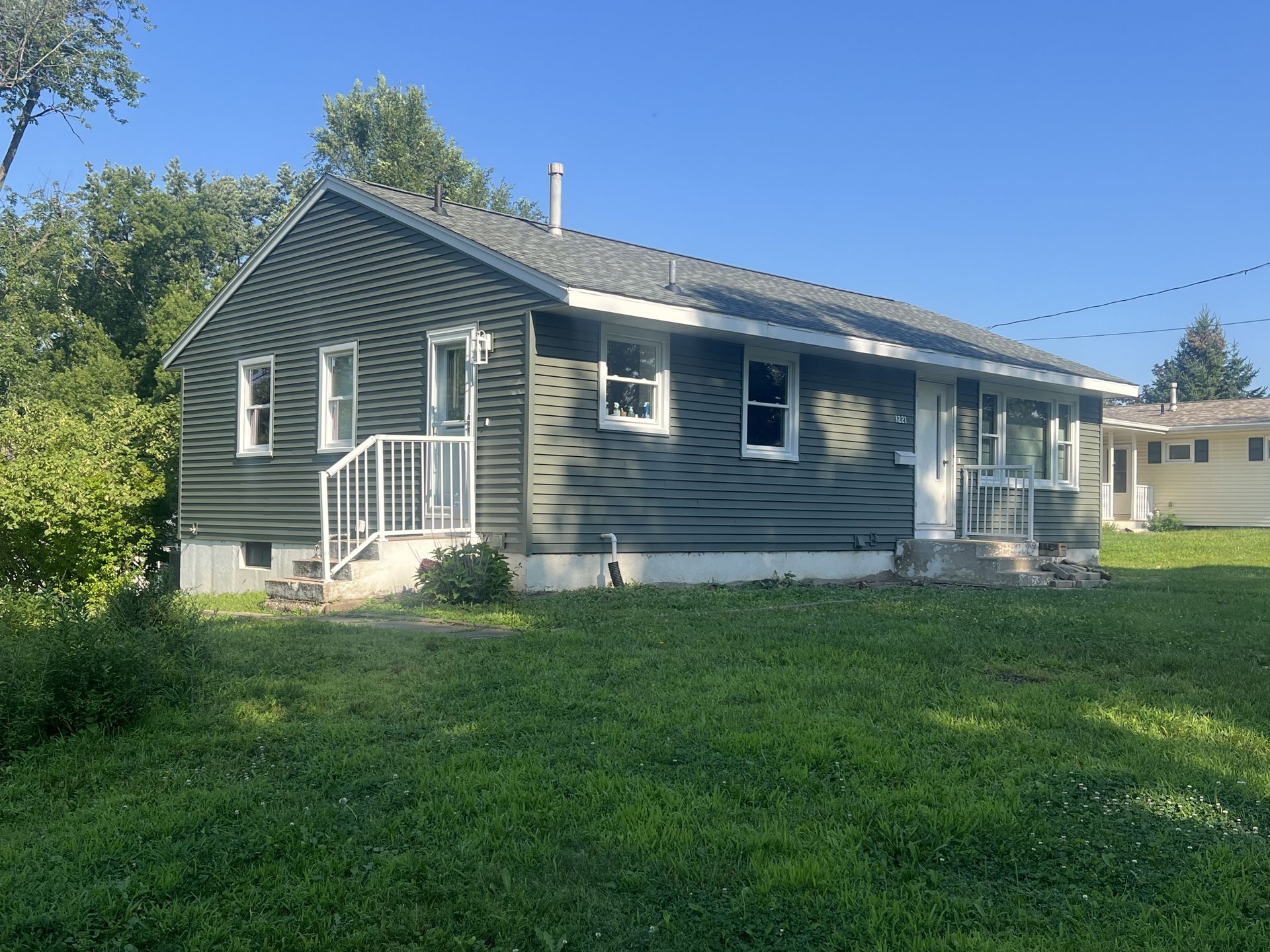 A single-story house with a white a gray blue siding and steps sits on a grassy lawn under a clear blue sky.