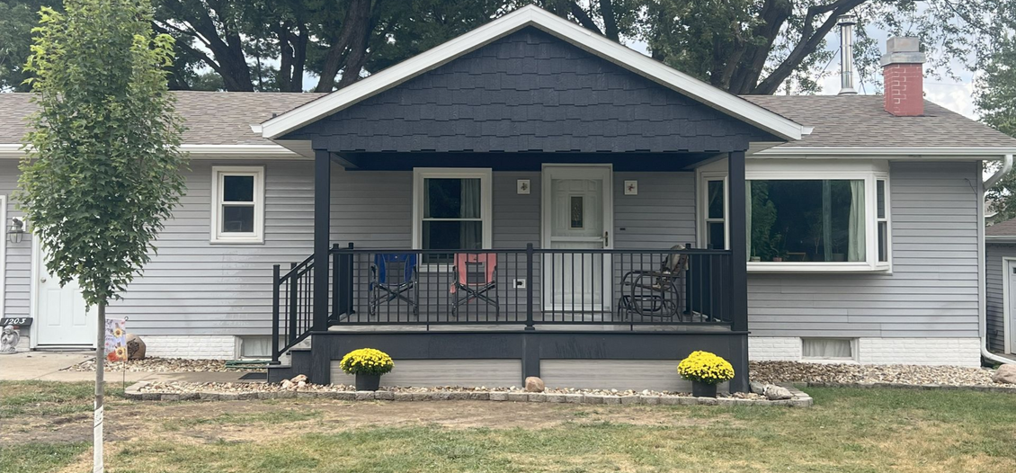 A gray house with a black deck and trim. Yellow flowers sit on the porch.