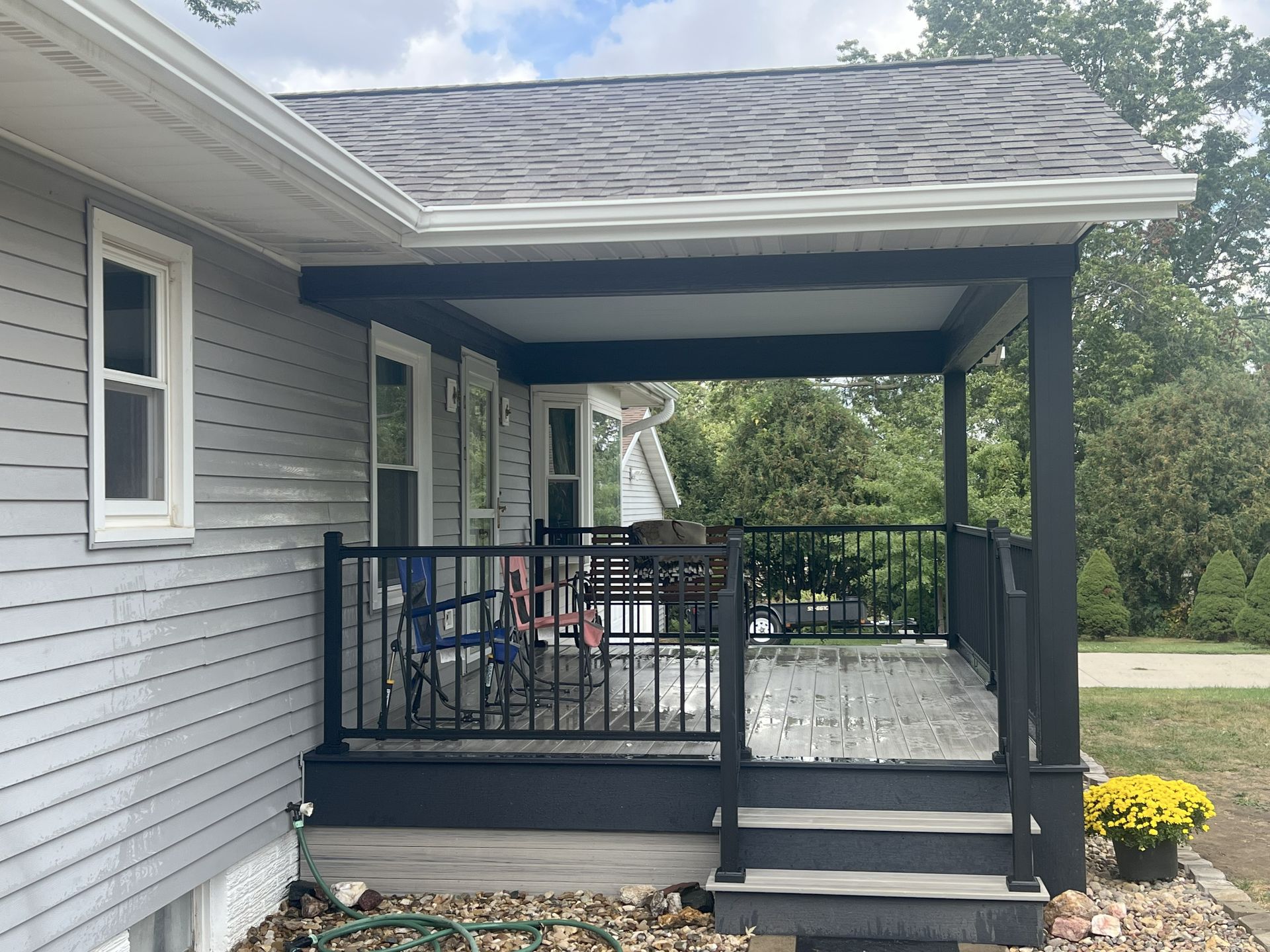 A small porch on a gray house, with black railings and a roof. Flowers sit in a pot.