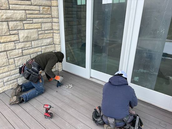 Two men are working on a deck next to a brick wall.