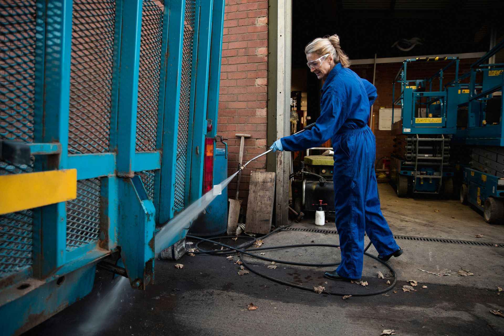 A woman in blue overalls is cleaning a blue fence with a high pressure washer.