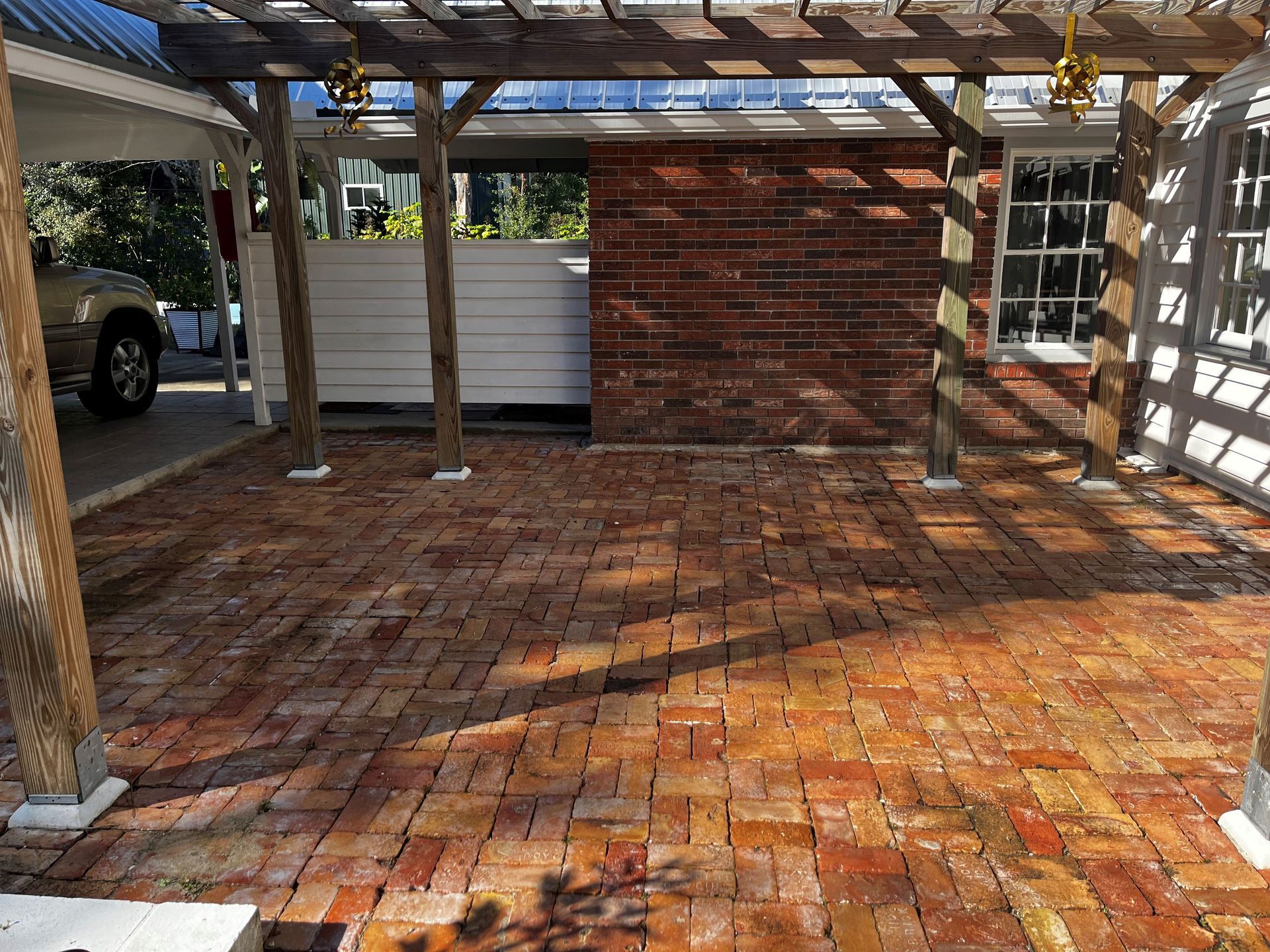 A car is parked under a pergola in a brick driveway.