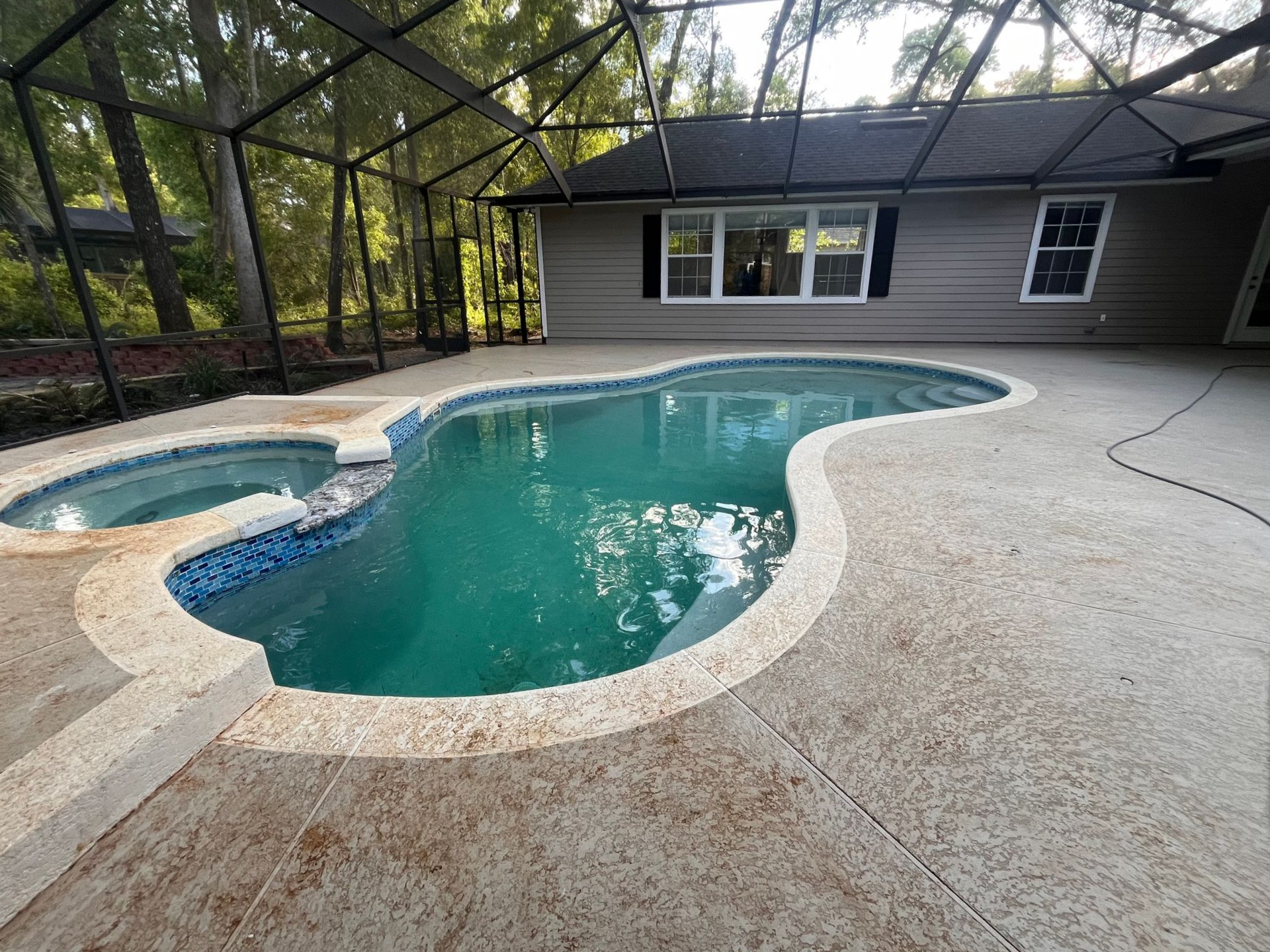 A pool with attached spa under a screened enclosure. Grey concrete surround. House in background.