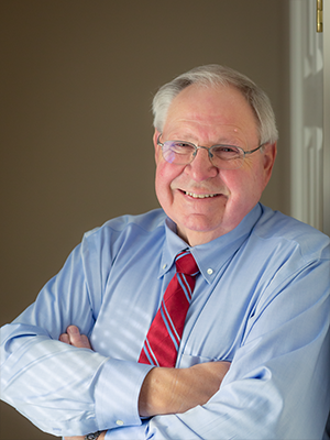 A man in a blue shirt and red tie is smiling with his arms crossed