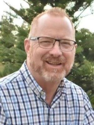A man with a beard and glasses is wearing a plaid shirt and smiling.