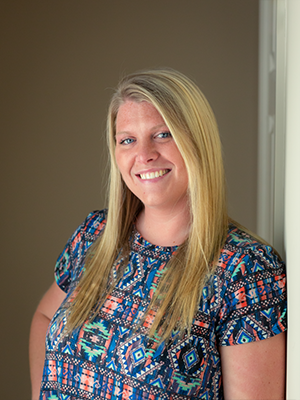 A woman in a colorful shirt is leaning against a wall and smiling.