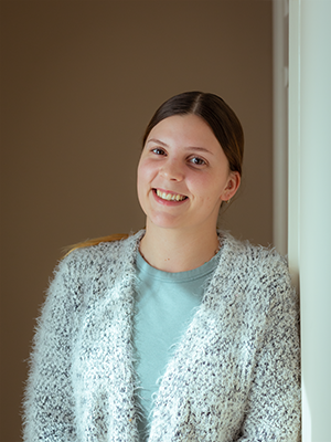 A woman in a sweater is leaning against a wall and smiling.
