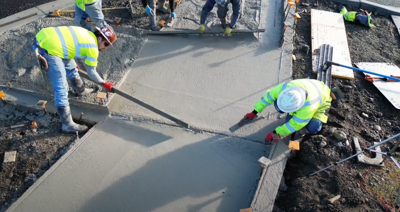 Person using a squeegee to spread wet, brown material on a concrete surface. Outdoor setting.