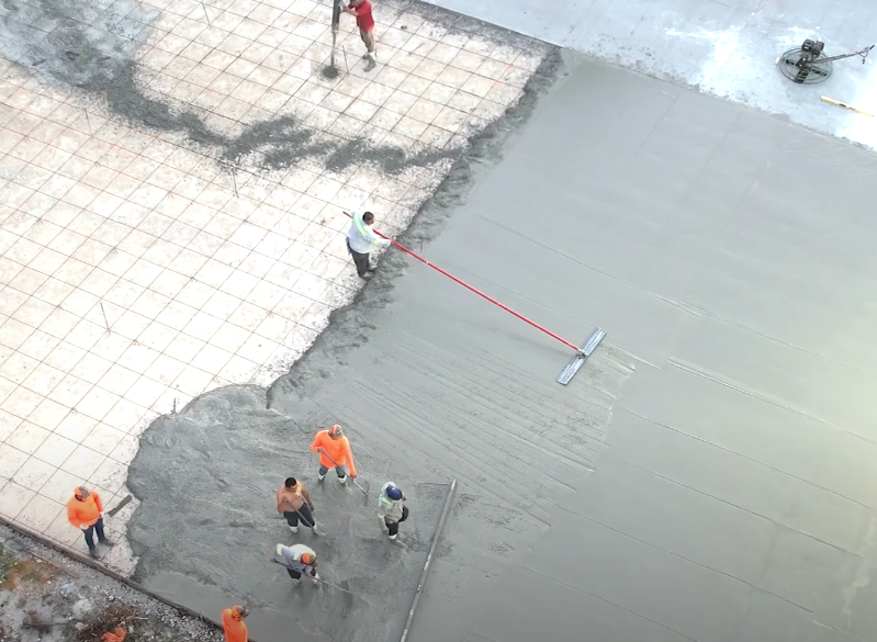 Construction workers smoothing wet concrete. Steel mesh visible. Bright colors: orange, red. Outdoors.