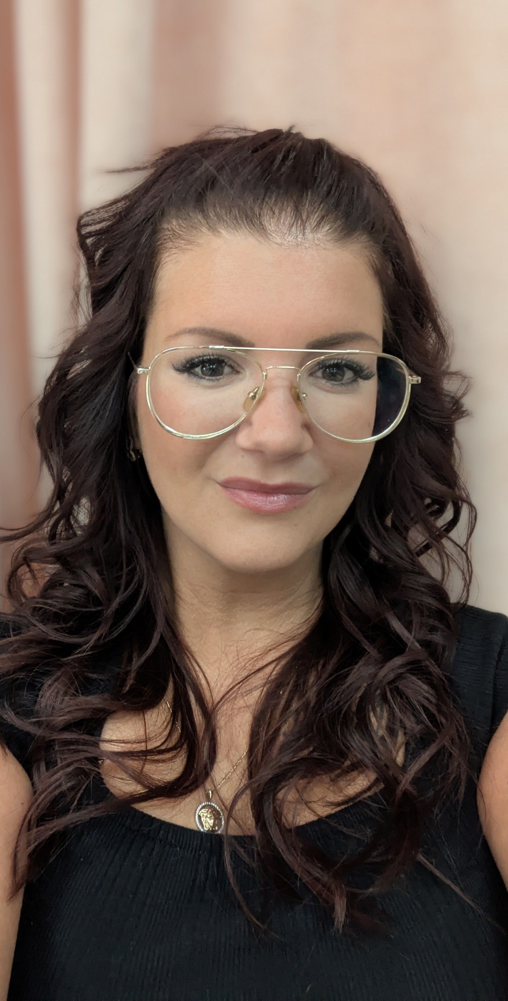 Une femme aux cheveux noirs bouclés, portant des lunettes à monture dorée, sourit à la caméra.