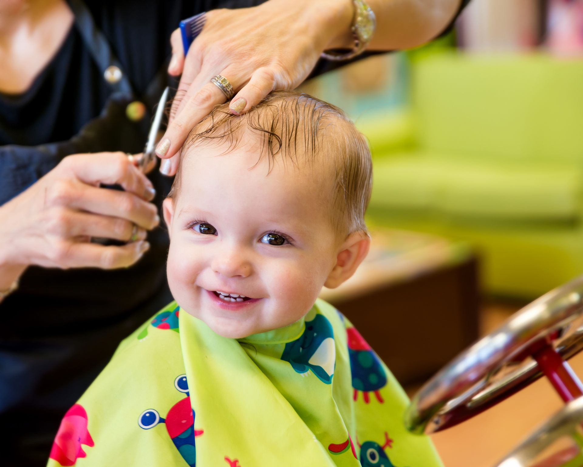 Un bébé souriant se fait couper les cheveux dans un salon de coiffure. Cape verte, ciseaux pour couper les cheveux, ambiance lumineuse.