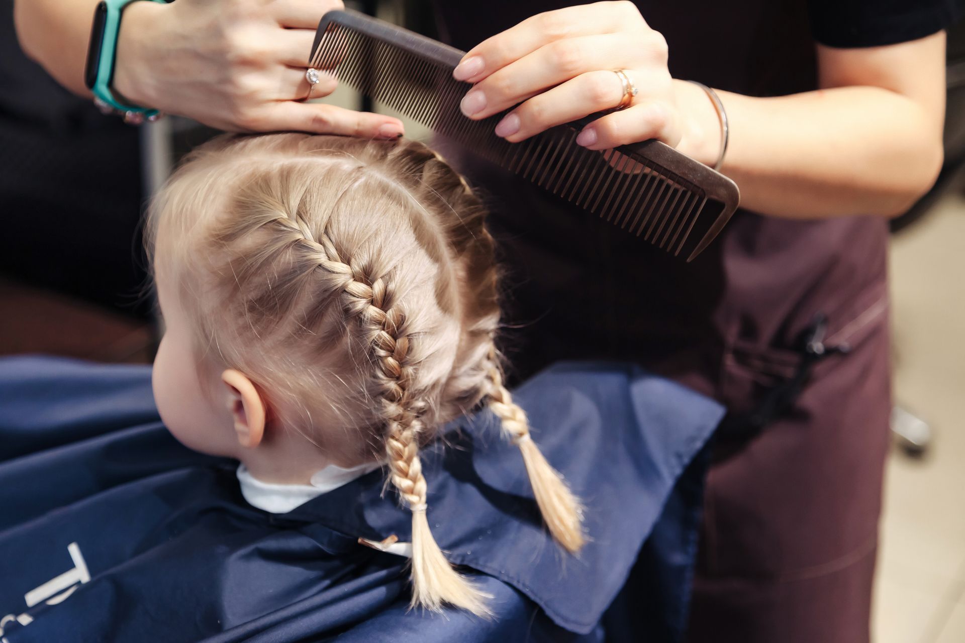 Une personne tresse les cheveux blonds d'une jeune fille en deux tresses dans un salon, la fille porte une cape bleue.