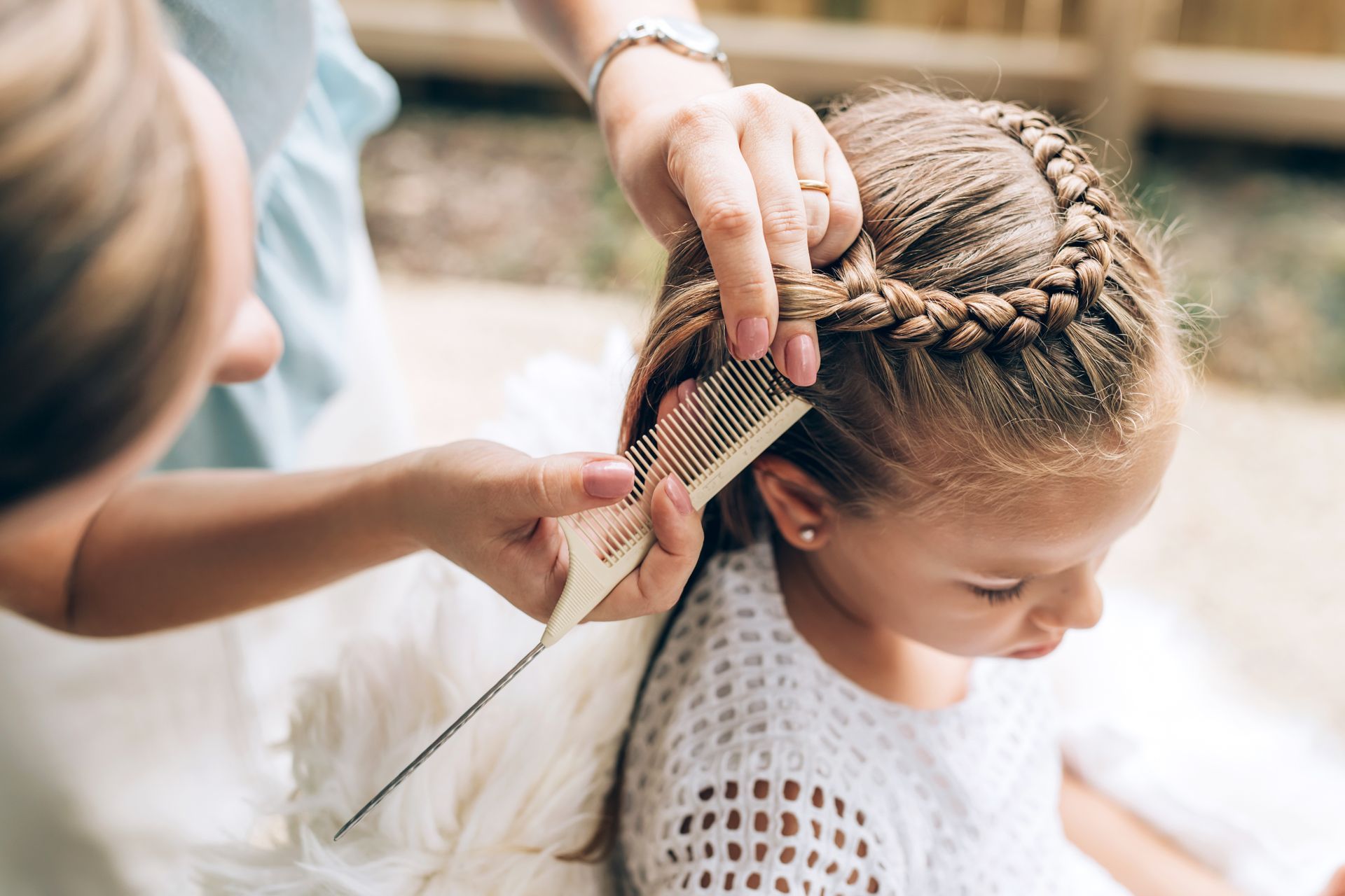 Une femme tresse les cheveux d'une jeune fille en extérieur. La fille regarde vers le bas tandis qu'une femme lui peigne et lui tresse les cheveux.