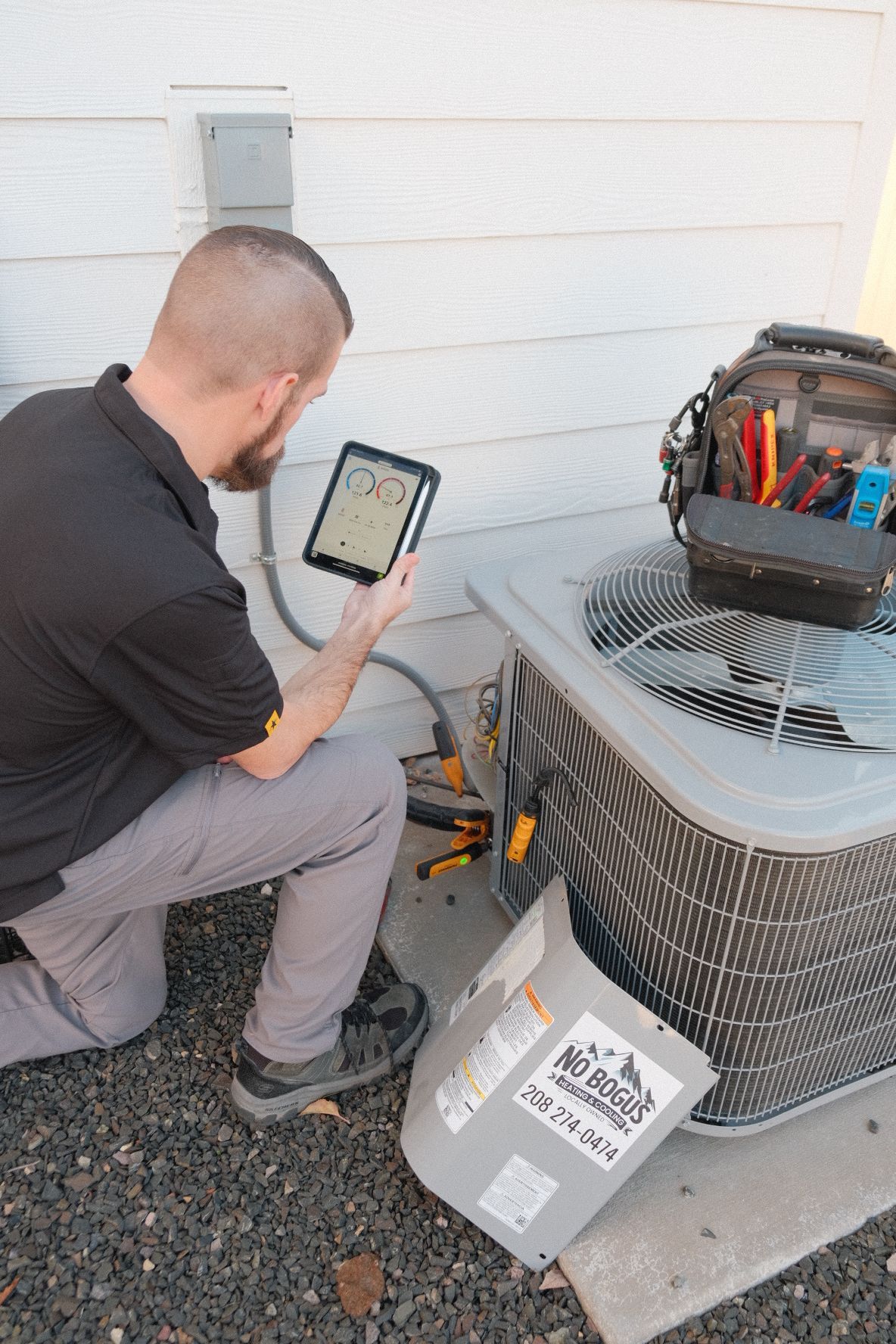 A man is kneeling down next to an air conditioner while using a tablet.