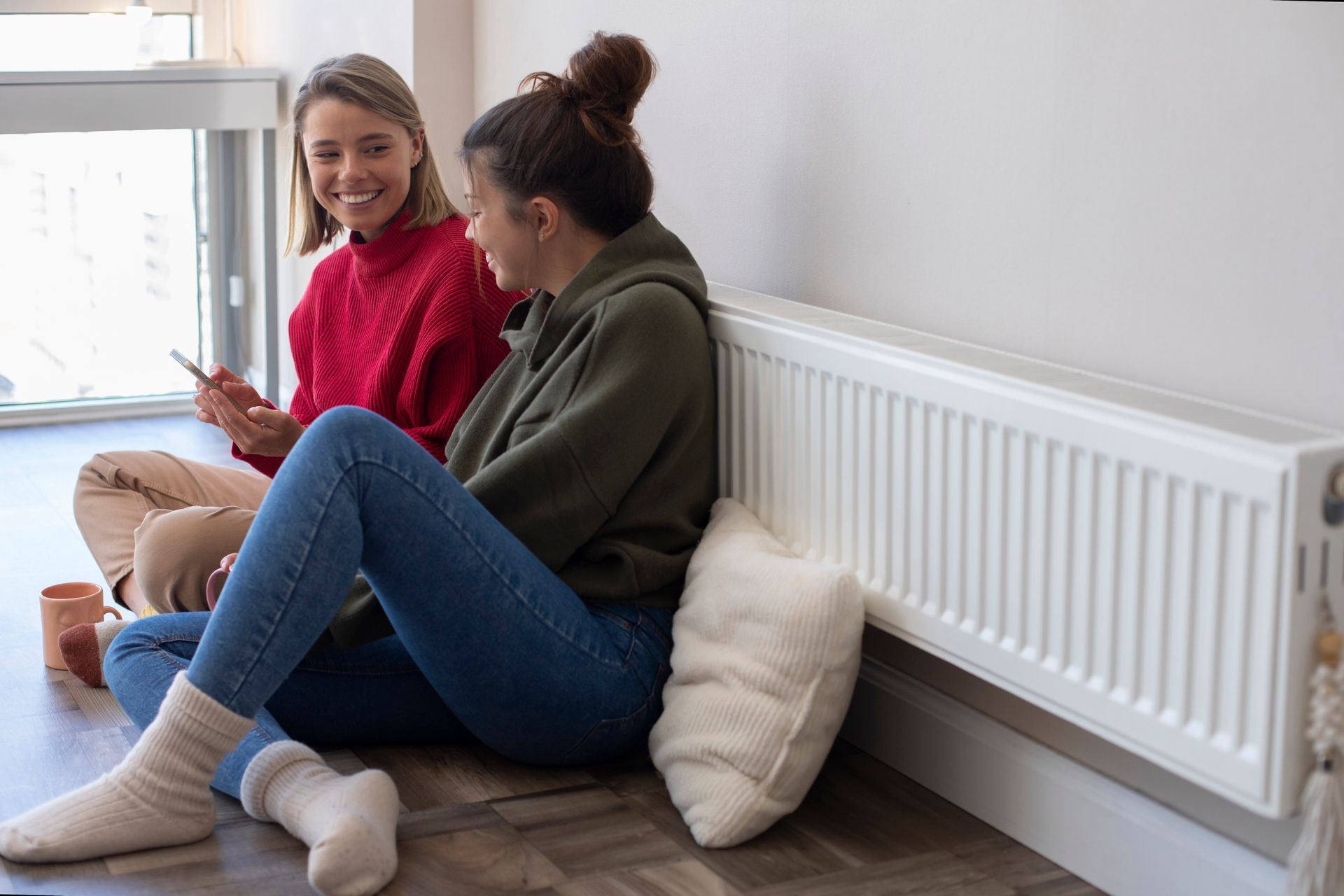 Two people sit on floor by a radiator, one looking at phone and smiling, the other listening and laughing.