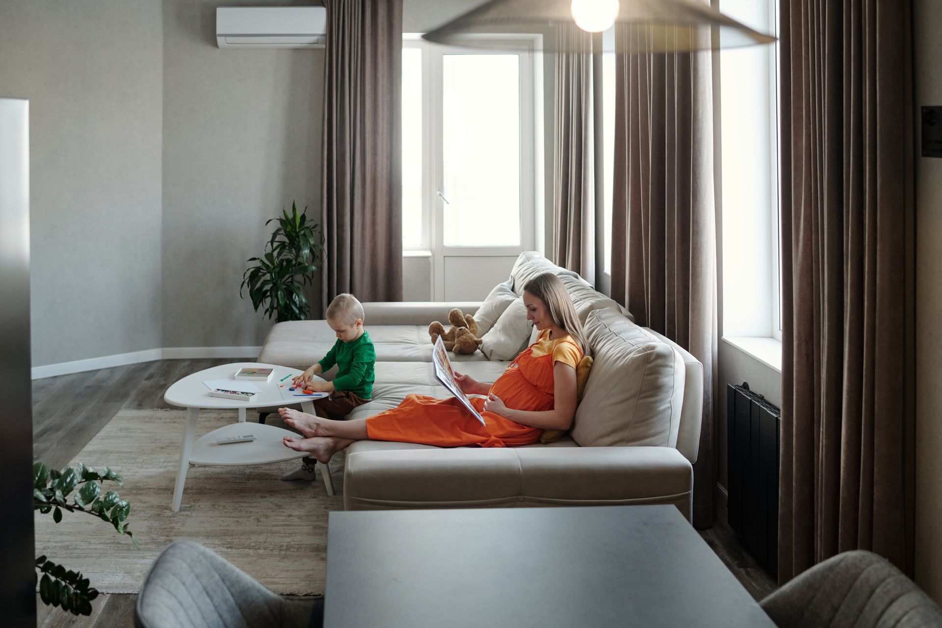 Woman reading book on couch with child in living room. Neutral colors.