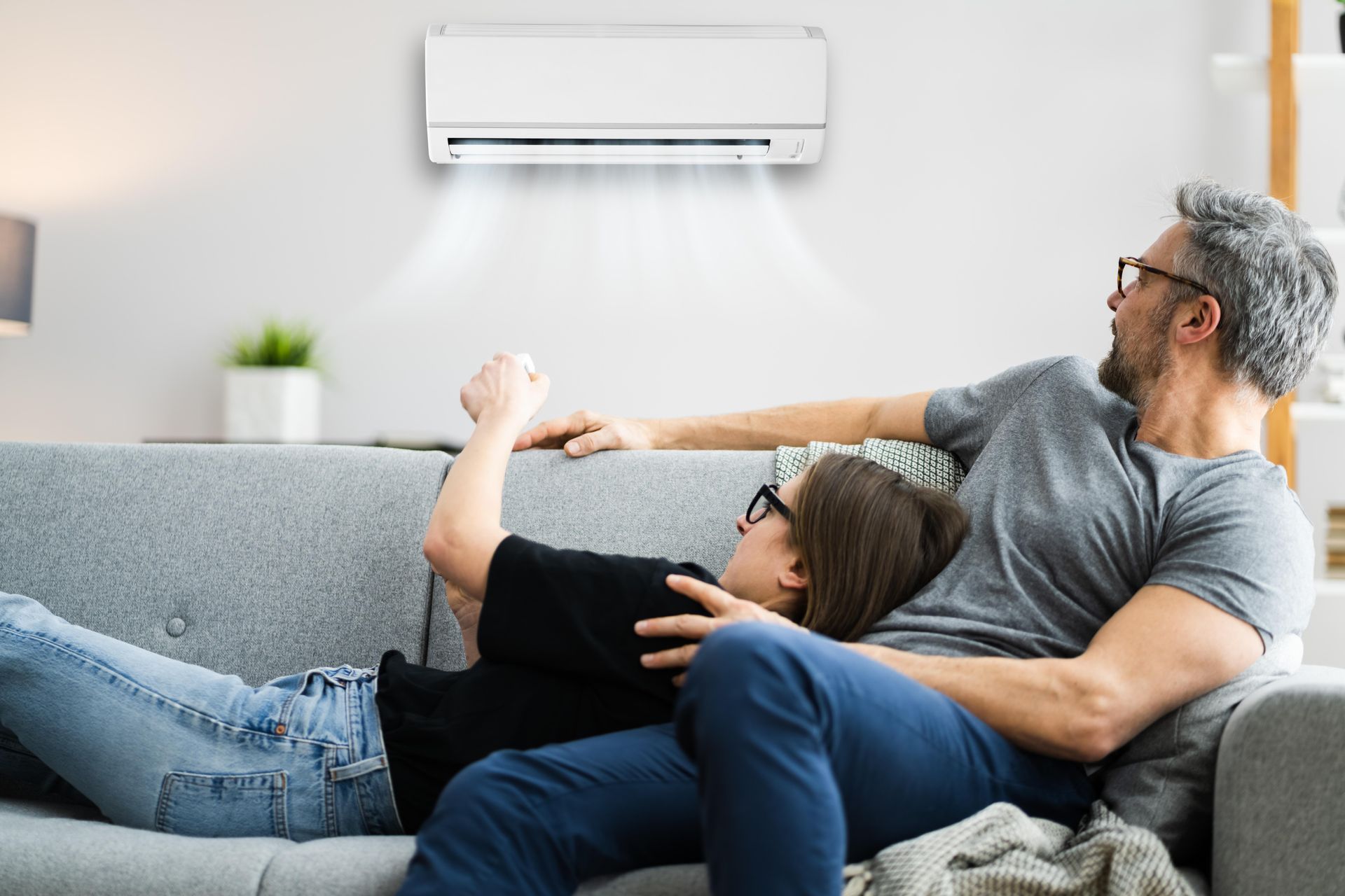 A man and a woman are laying on a couch under an air conditioner.