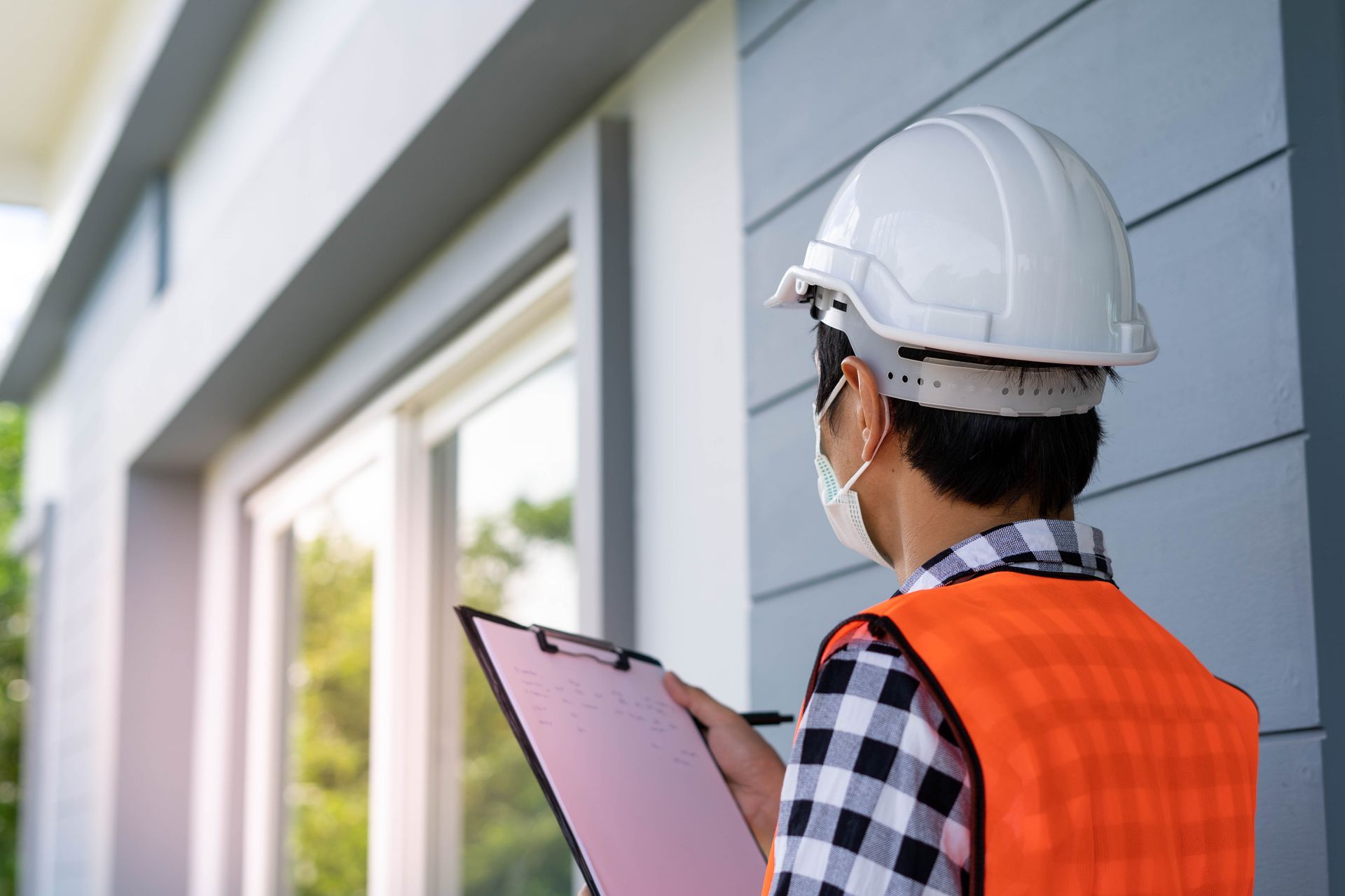 A man wearing a hard hat and safety vest is holding a clipboard in front of a building.