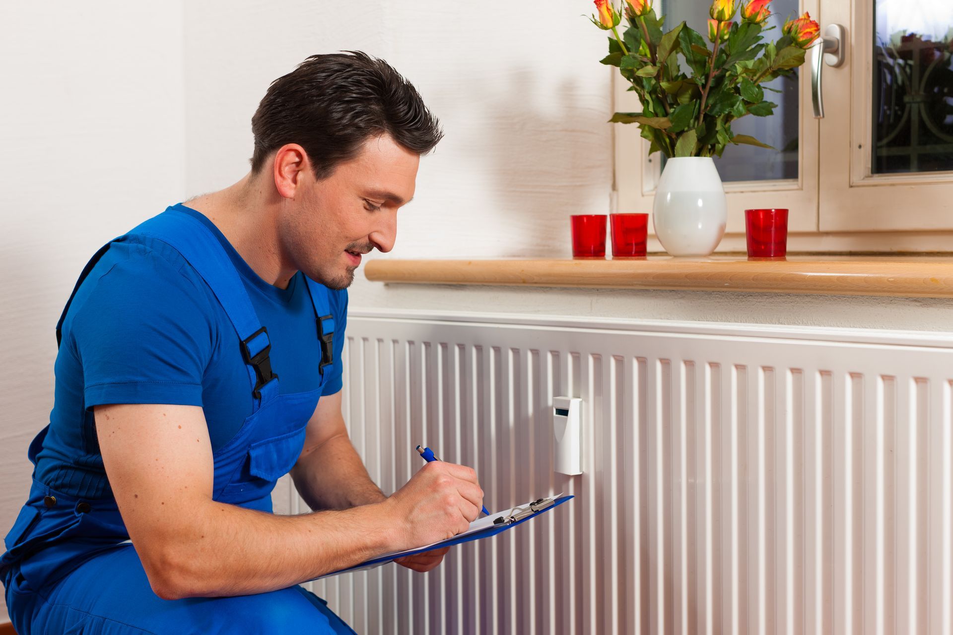 A man is kneeling down in front of a radiator and writing on a clipboard.