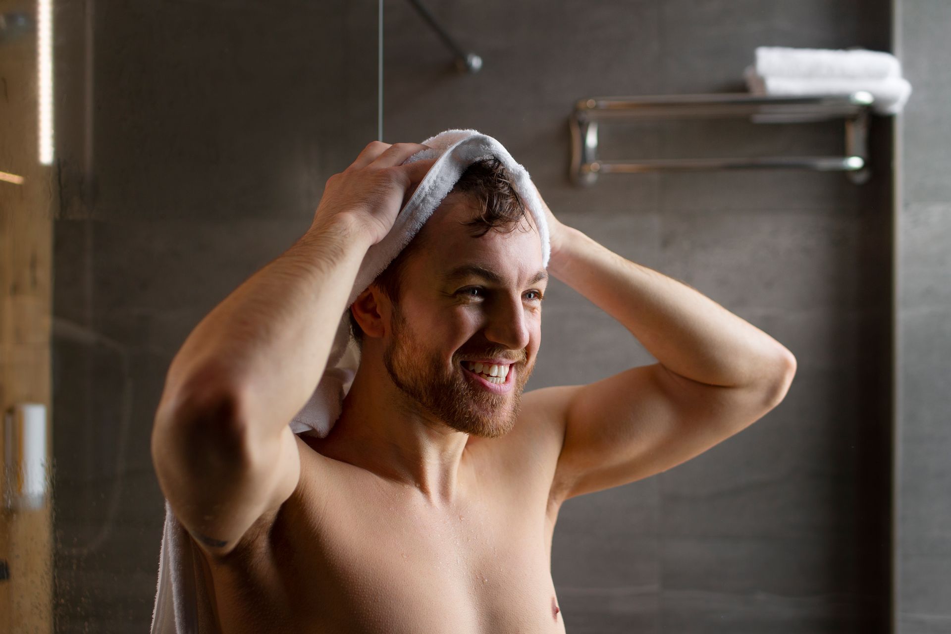 A shirtless man is wrapping a towel around his head in a bathroom.