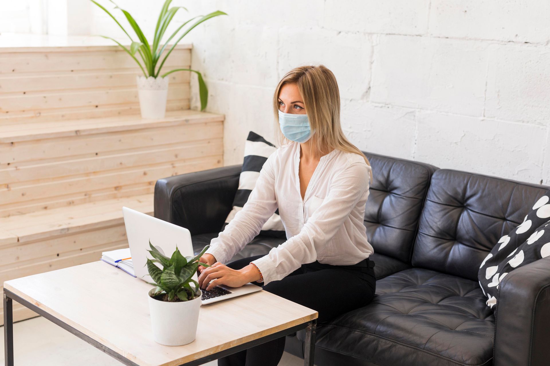 A woman wearing a mask is sitting on a couch using a laptop computer.