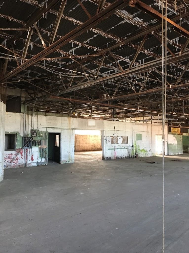 Interior view of a large, dilapidated warehouse. Concrete floor, metal roof structure, and open doorways.