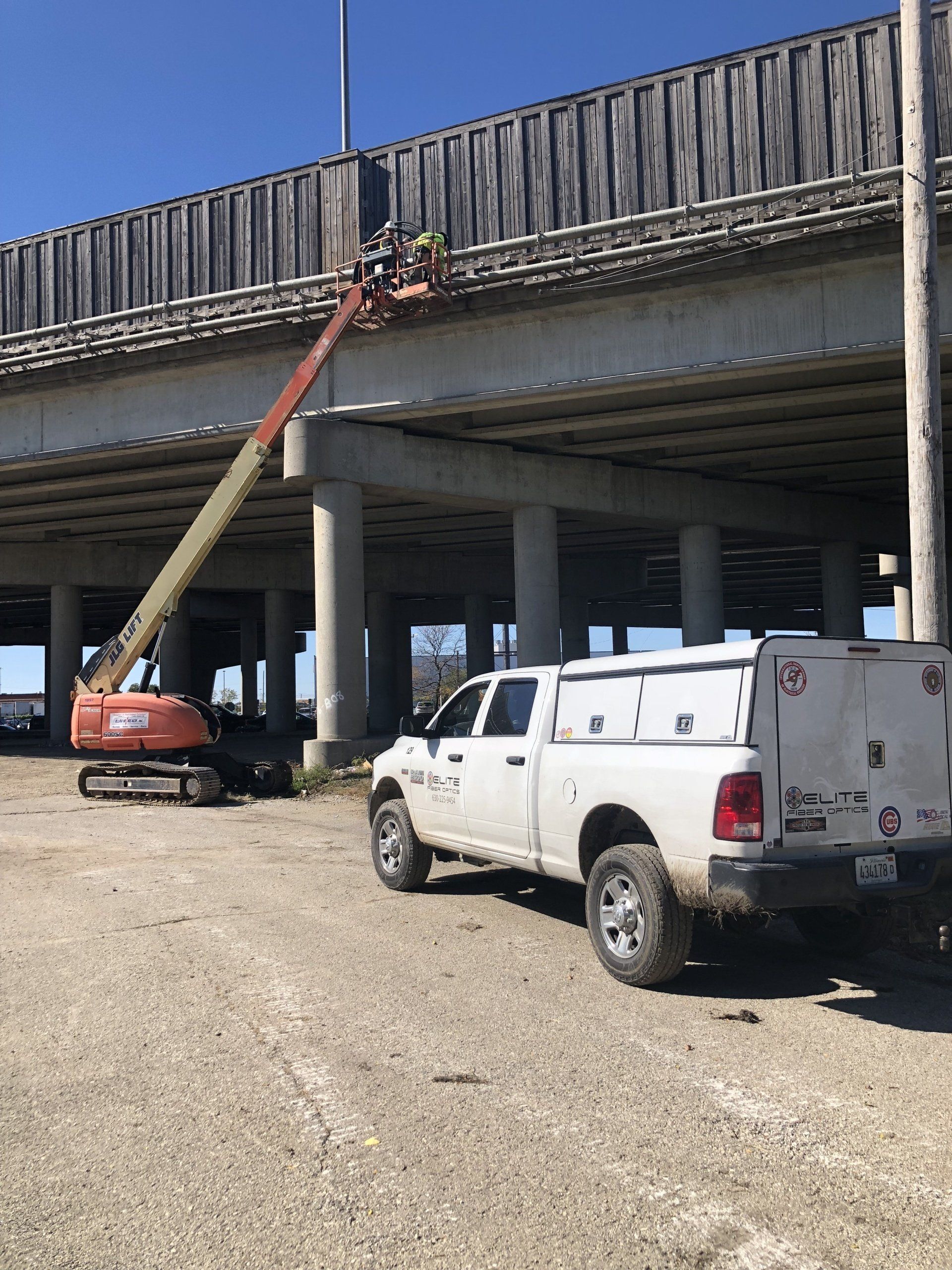 A white truck is parked under a bridge with a crane in the background.