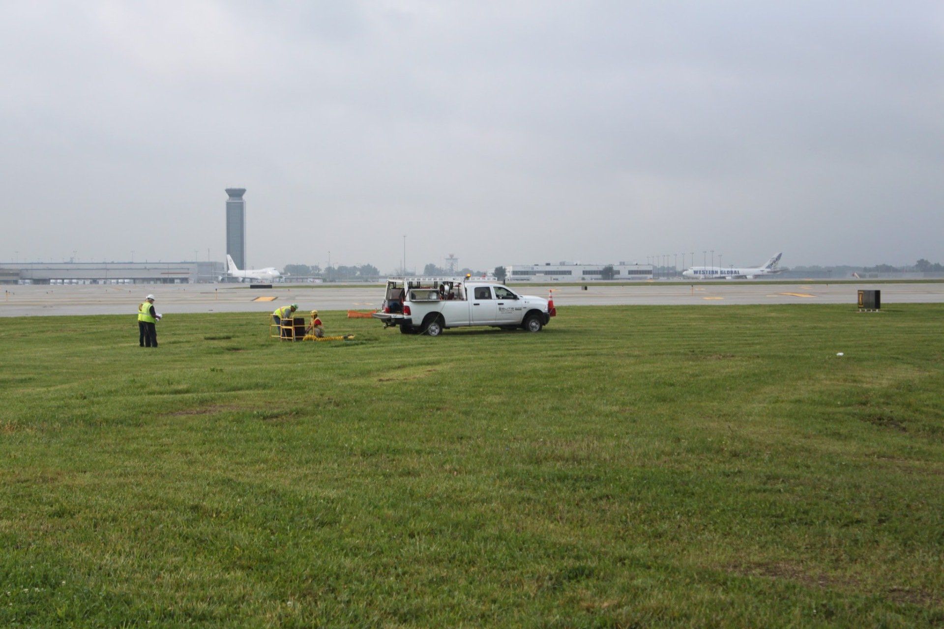 A white truck is parked in a grassy field next to an airport runway.