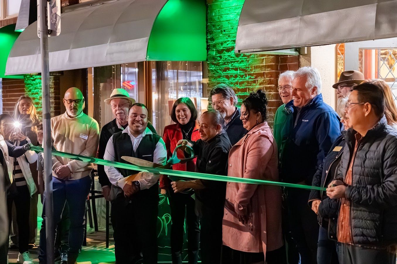 A group of people are standing in front of a building cutting a green ribbon.