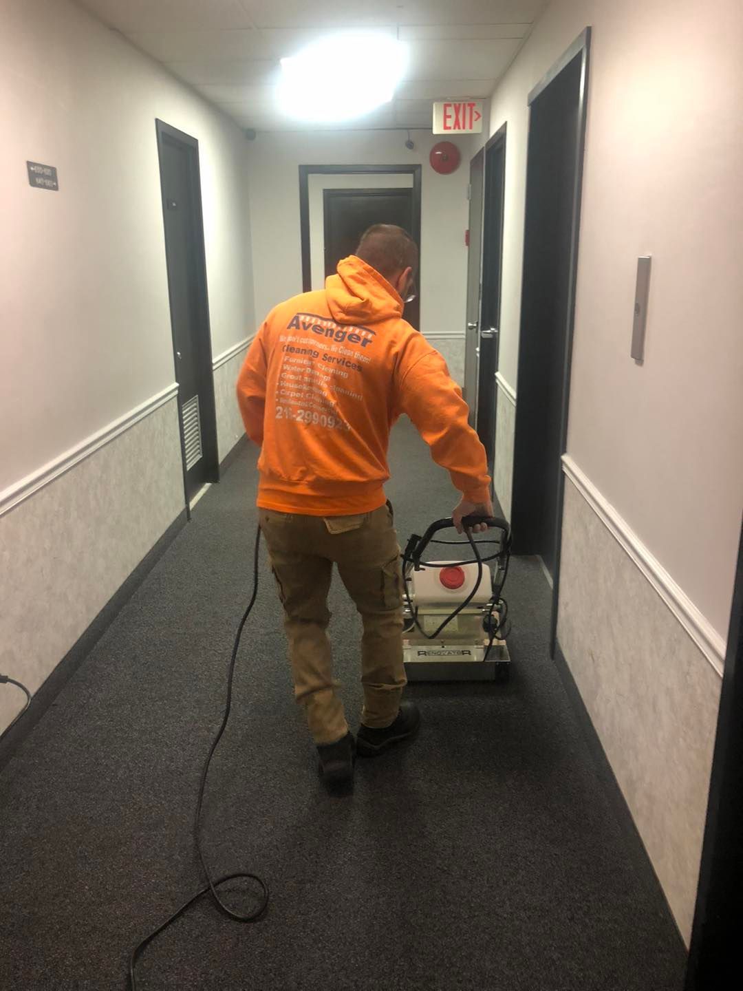 A man in an orange hoodie is cleaning a hallway with a vacuum cleaner.