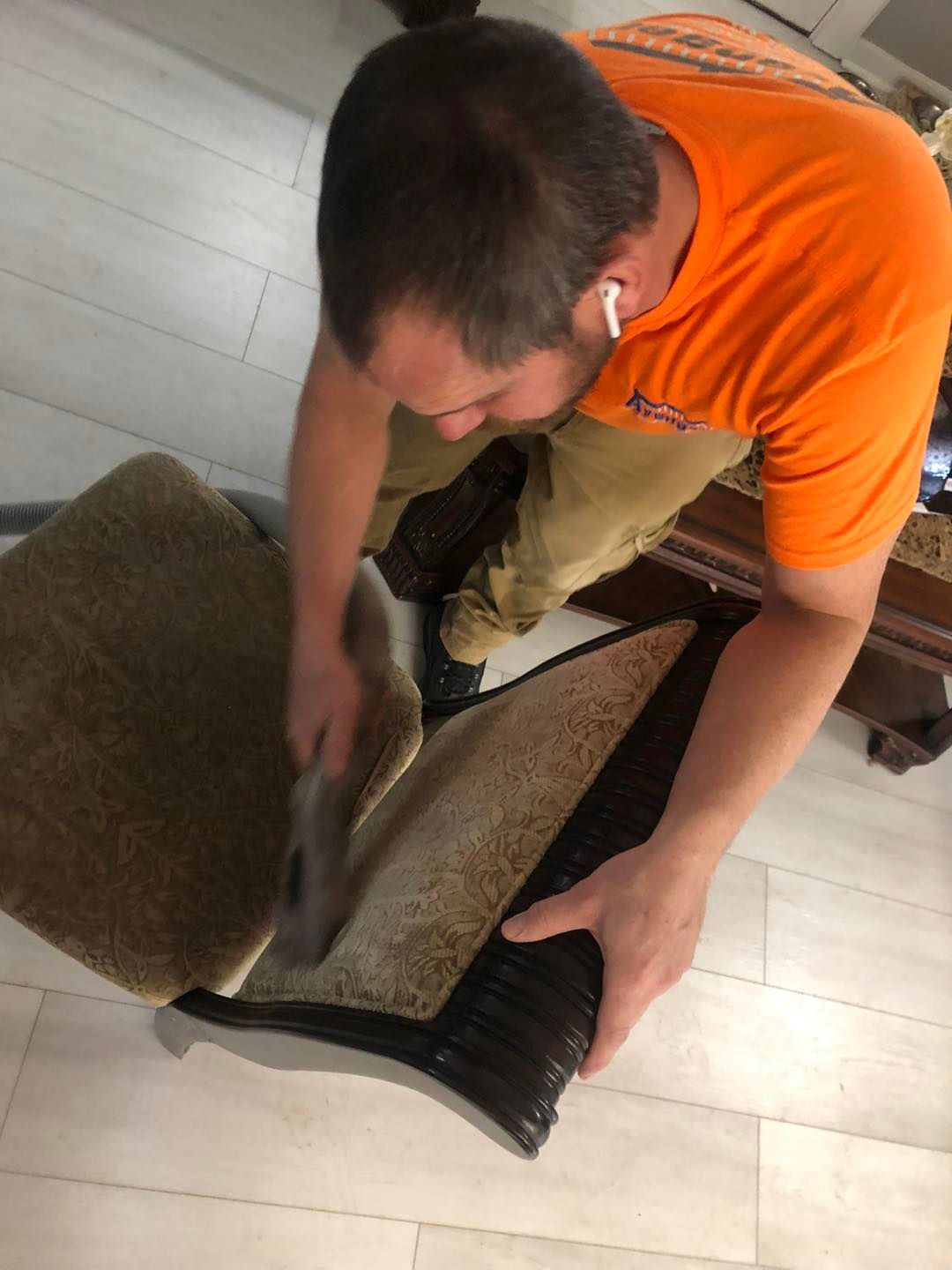 A man in an orange shirt is cleaning a chair with a vacuum cleaner.