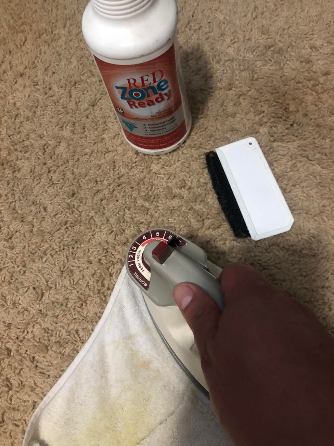A person is ironing a towel on a carpet next to a bottle of red zone ready.