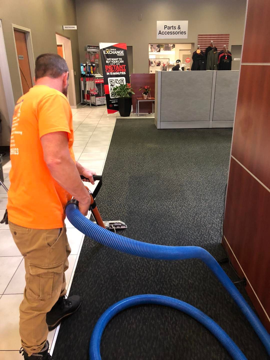 A man in an orange shirt is cleaning a carpet with a vacuum cleaner.