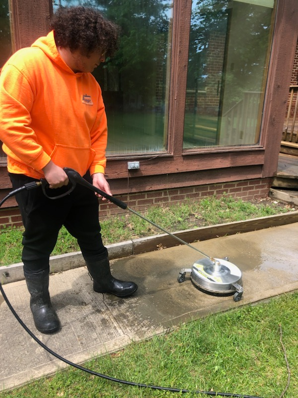 A man in an orange hoodie is cleaning a sidewalk with a pressure washer.