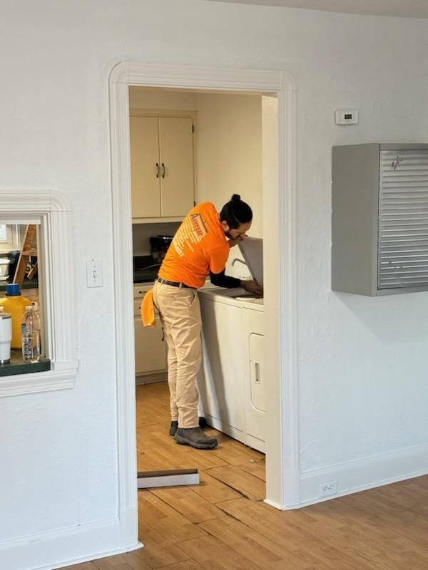 A man in an orange shirt is working on a washing machine in a kitchen