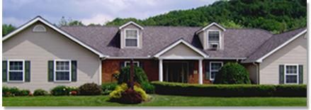 A beige ranch-style house with a dark gray roof, green lawn, and shrubbery in front of a wooded mountain.