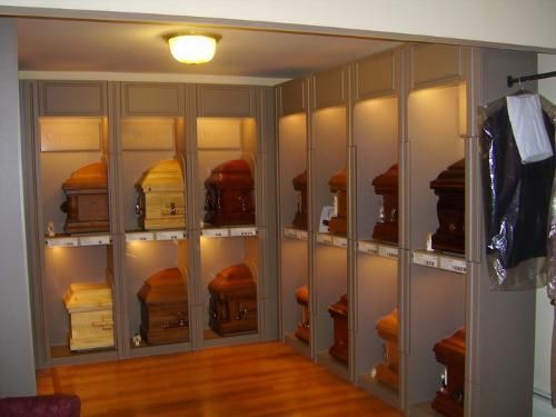 Room displaying caskets in individual lit alcoves. Wooden floor, walls, and lighting. A garment bag hangs on a rod.