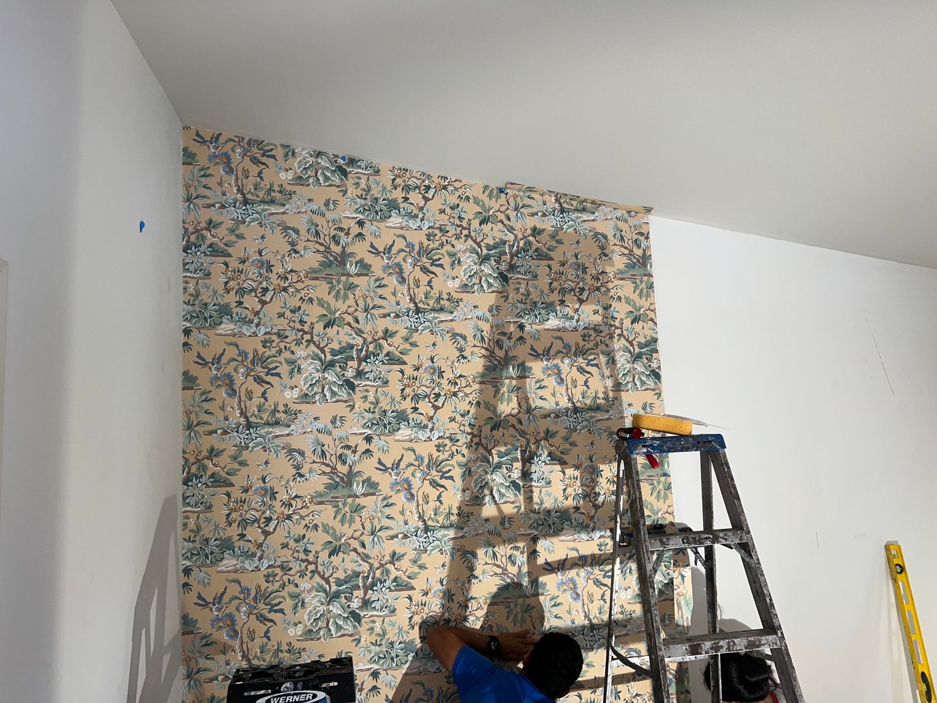A man is standing on a ladder installing wallpaper on a wall.