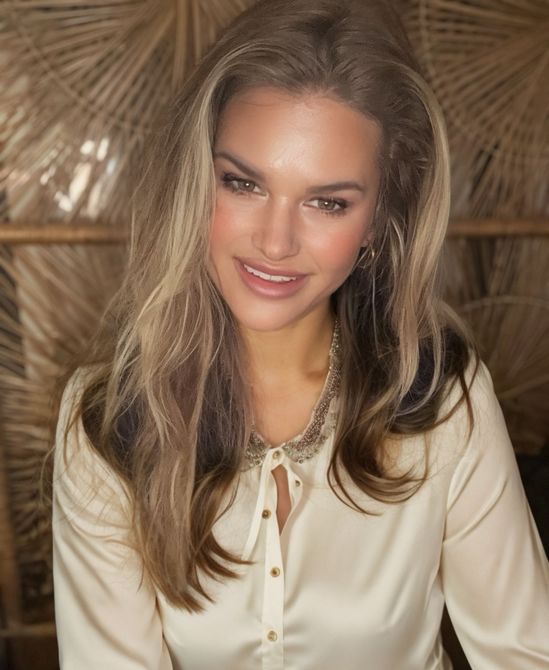 Blonde woman smiling, wearing a cream-colored satin blouse, against a woven, light-brown background.