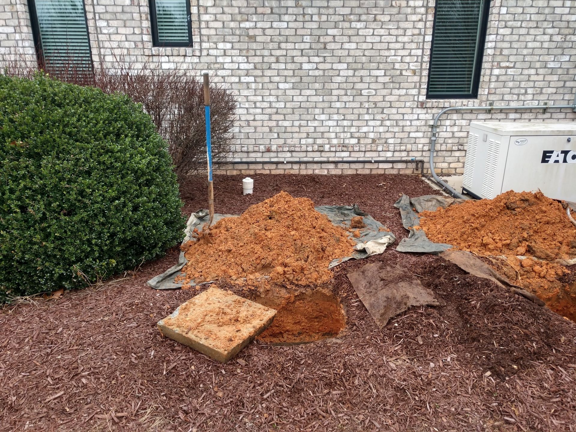 A yard with red mulch, piles of dirt, a bush, and a generator next to a brick building.