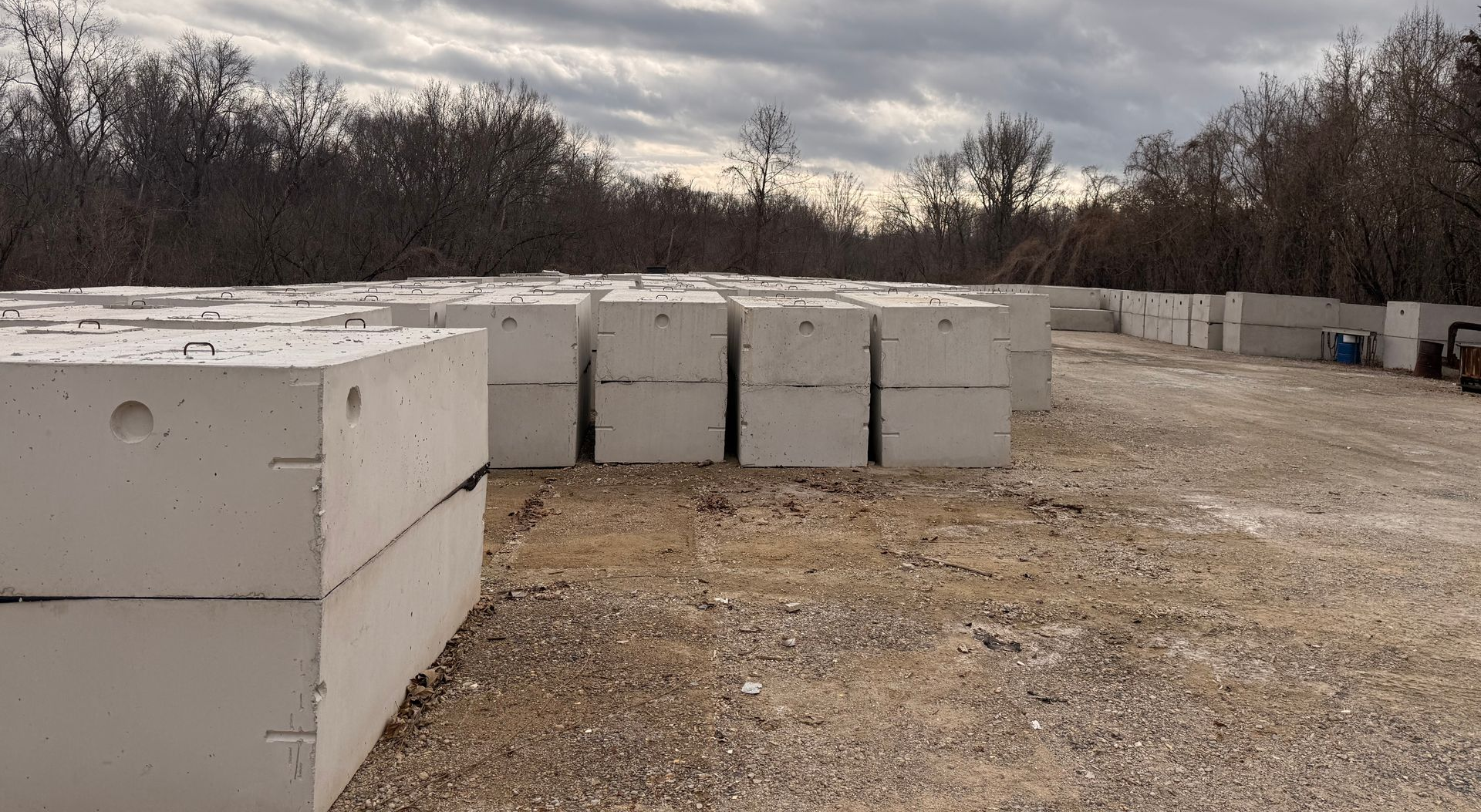 Concrete blocks stacked outdoors, possibly at a construction site. Gray, gravel ground, bare trees in the background.