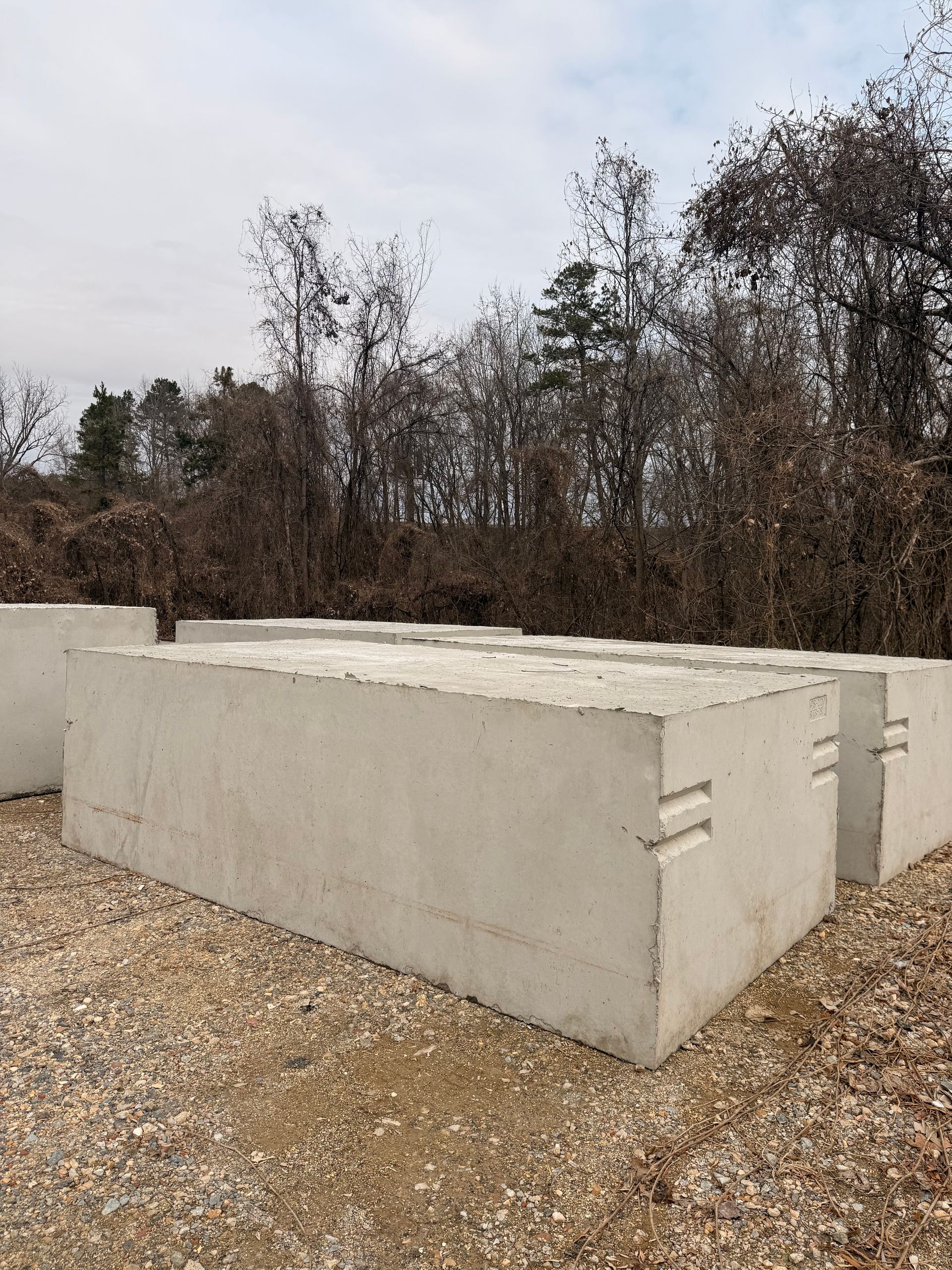 Gray concrete blocks stacked outdoors on gravel, with trees in the background.