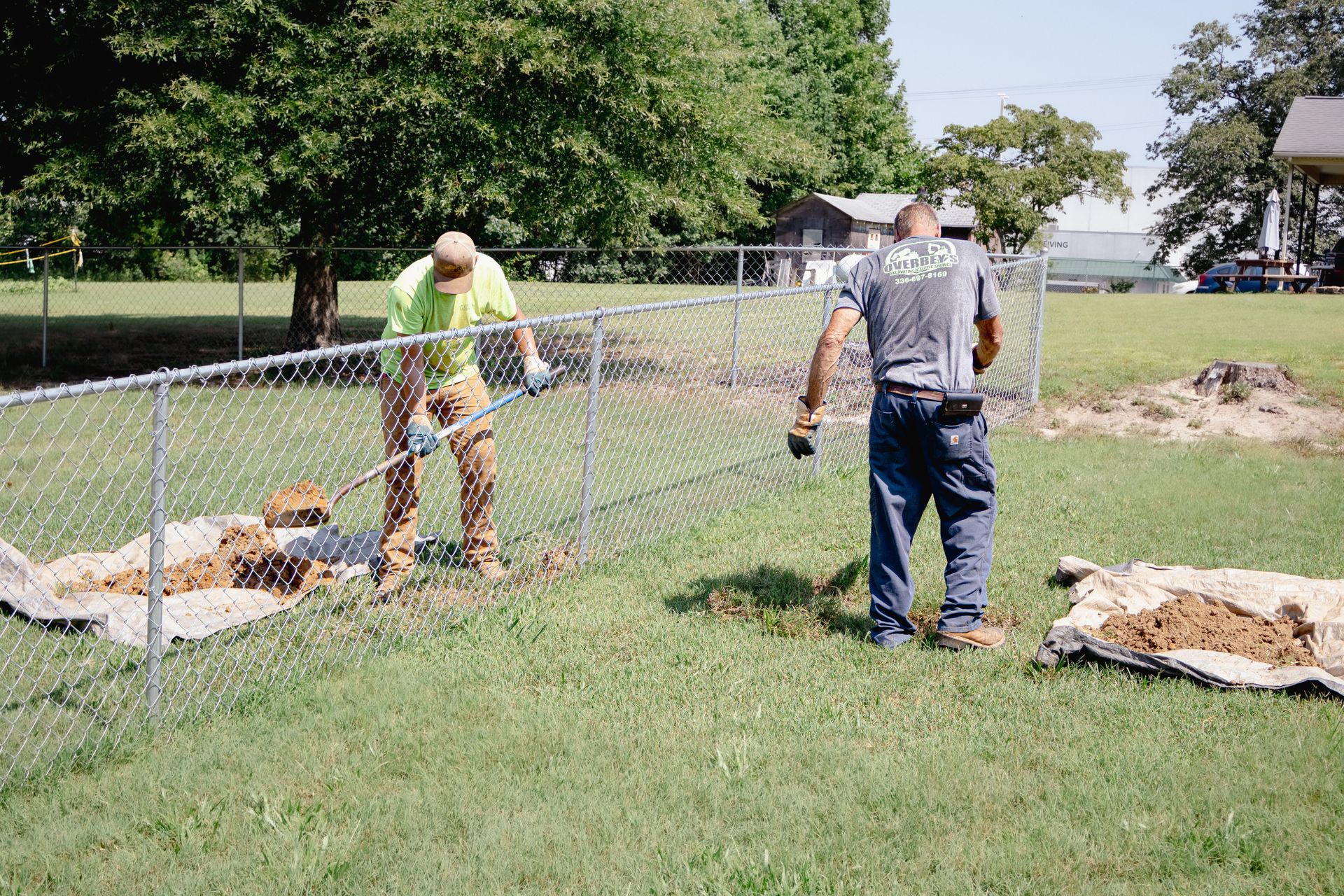 Two workers installing a chain-link fence on a grassy lawn. One digs while the other assists.
